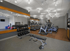 A modern gym with treadmills, ellipticals, dumbbells, a weight bench, and mirrors. Walls are gray and orange, and the flooring is a mix of carpet and wood.