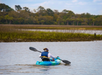 A person wearing a life jacket kayaks on a calm body of water with marsh grass and trees in the background.