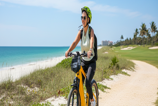 A woman wearing a helmet and sunglasses rides a yellow bicycle on a sandy path by the beach, with grass, palm trees, and blue sky in the background.