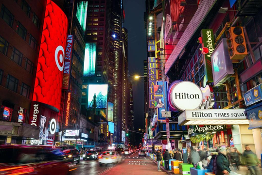 Busy Times Square street scene at night with illuminated signs, including Target, Hilton, and Applebee's, and blurred motion of cars and pedestrians.