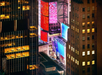 Aerial view of illuminated billboards and buildings at night between high-rise structures in a cityscape.