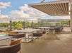 Outdoor rooftop dining area with modern tables and chairs, glass railing, and a view of trees and a pool below under a partly cloudy sky.