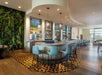 Modern hotel bar with curved counter, blue chairs, three TV screens, hanging lights, patterned floor, and a green plant wall feature on the left side.