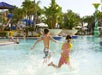 Two children run and splash in a large outdoor swimming pool surrounded by palm trees, lounge chairs, and water slides on a sunny day.