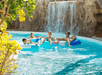 Four people are floating on inner tubes in a pool near a waterfall, surrounded by rocks and greenery.