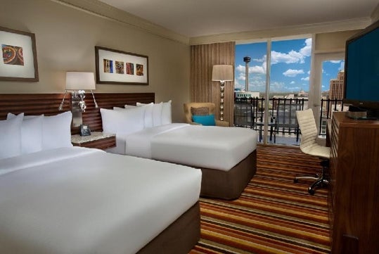 Hotel room with two double beds, a desk and chair, a TV, artwork on the wall, and a balcony view of city buildings under a blue sky with clouds.