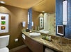 Modern hotel bathroom with granite countertop, vessel sink, large mirror, blue tile accents, toiletries, towel rack, wall art, and a shower with a curtain and grab bars.