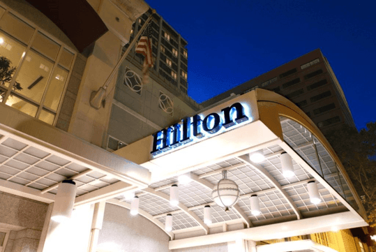 The front entrance to the Hilton Portland Downtown at night with lights illuminating the covered entrance and a glowing Hilton sign.