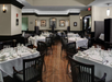 A formal restaurant dining area with neatly set tables, white tablecloths, black chairs, and framed artwork on cream-colored walls. The room has wood flooring and a modern, elegant atmosphere.