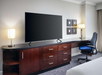 Hotel room with a large flat-screen TV on a wooden desk, flanked by two lamps. There's a blue office chair and abstract wall art above the desk.