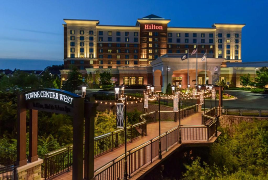 Exterior of a Hilton hotel at dusk, with a lighted pathway and bridge in the foreground.