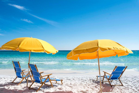Two yellow beach umbrellas and four blue chairs on a sandy beach by the blue ocean under a clear sky.