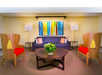 A living room with a blue sofa, colorful abstract painting, wooden chairs with red cushions, and a round coffee table with a floral centerpiece. Two lamps flank the sofa against a patterned wall.