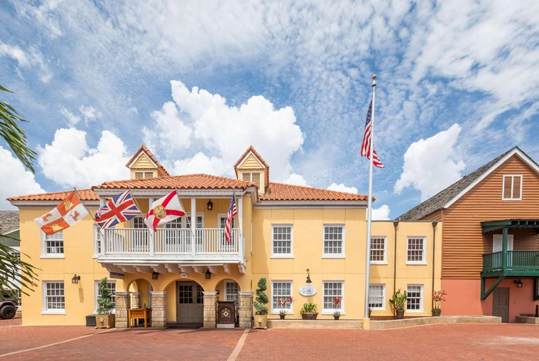 A charming exterior of the hotel featuring classic Spanish-style architecture with warm yellow walls, a red-tiled roof, and a row of international flags waving above the entrance, all set against a bright blue sky with fluffy clouds.