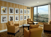 A cozy seating area in a hotel room with four brown armchairs, two round tables, framed black-and-white photos on a patterned wall, and a large window with a view of an airport.