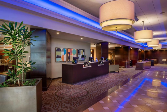 Hotel lobby with modern decor, featuring a reception desk, large ceiling lights, abstract artwork, and a potted plant.