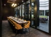 A long dining table with yellow chairs is set up in a modern, dimly lit restaurant room with reflective glass walls.