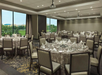 A banquet room with round tables set for dining, featuring white tablecloths, napkins, and glassware. Large windows provide a view of greenery outside.