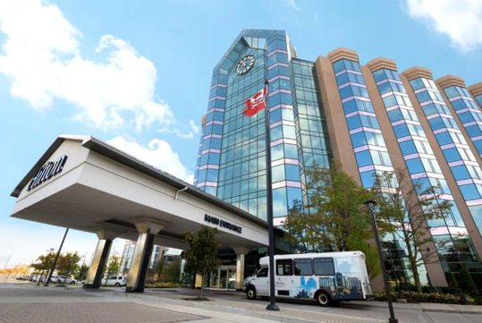 Modern building with a clock tower and large windows, featuring a covered entrance with a sign reading “Hilton” and a shuttle bus parked in front. A flag flutters beside the structure.