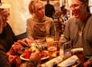 Four adults sit around a restaurant table with food and drinks, engaged in conversation. One person serves nachos from a shared plate.