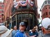 A group of people stands outside The Bell In Hand Tavern, a historic brick building with patriotic bunting decorations in an urban setting.