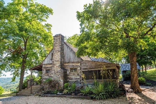 A rustic log cabin with a stone chimney sits among green trees and shrubs on a sunny day, with a gravel path in the foreground.