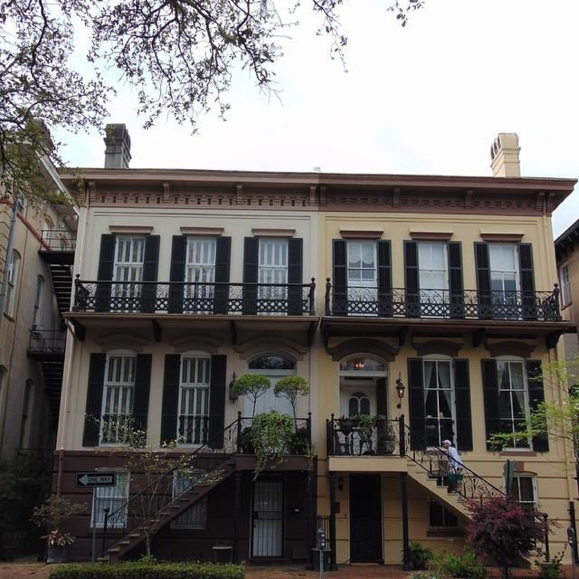 Historic two-story building with a symmetrical facade, wrought-iron balconies, and a staircase leading to the entrance. Trees frame the scene.