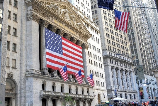 The facade of the New York Stock Exchange displays several large American flags in front of a historic building on Wall Street.
