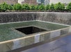 A large square reflecting pool with cascading water at the National September 11 Memorial, with trees and buildings in the background.