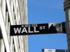 Black and white Wall Street street sign with building facade in background and blue sky above.