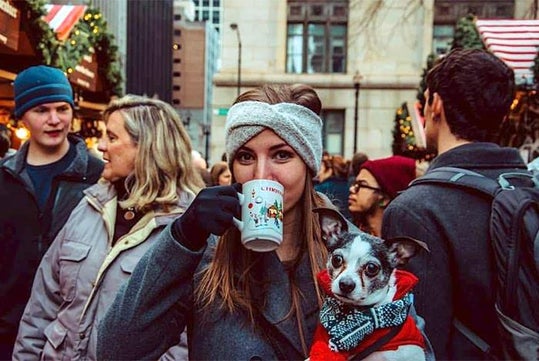 A woman in winter clothing drinks from a holiday mug and holds a small dog in a scarf, surrounded by people at an outdoor market.