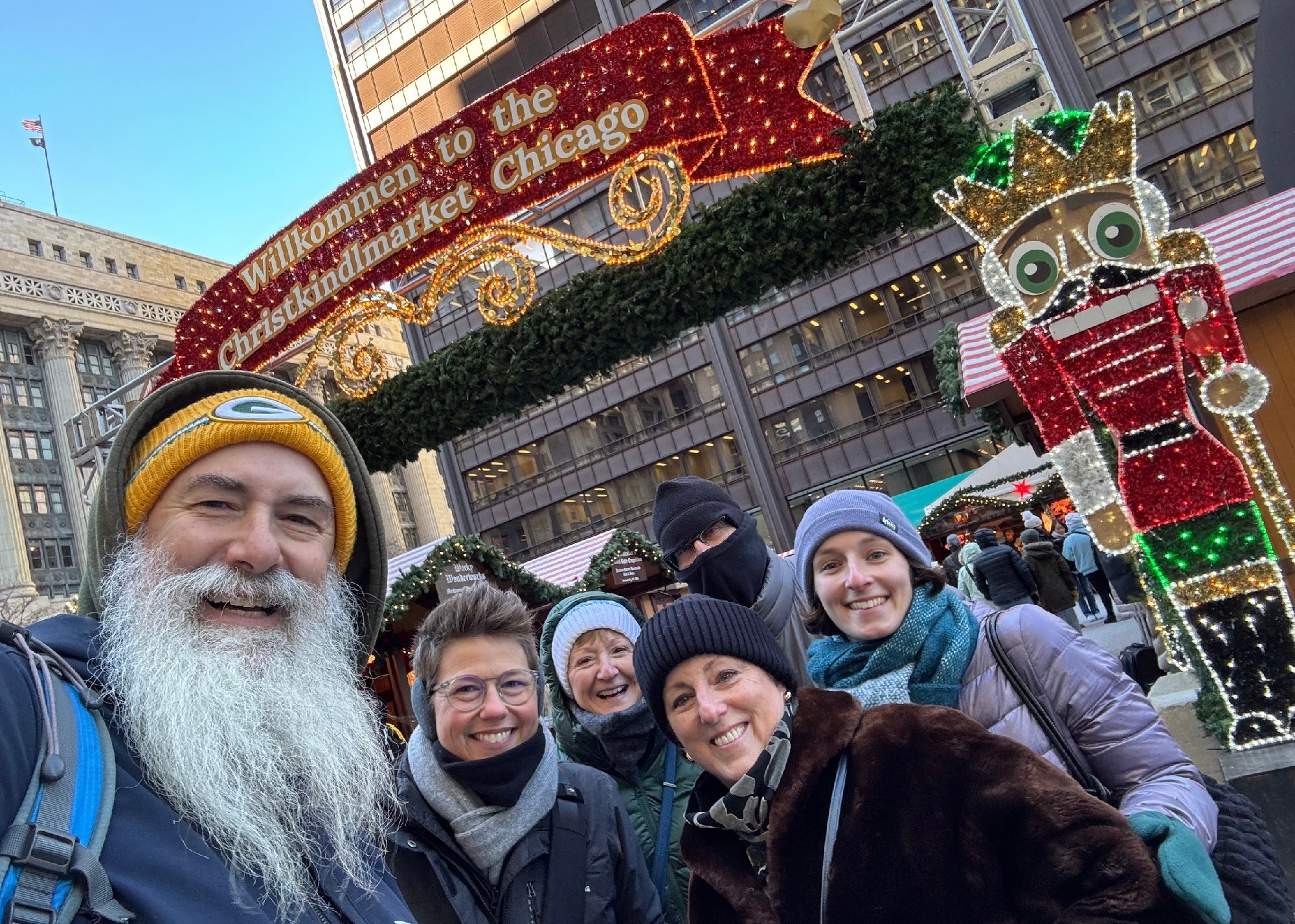 A group of six people smiles for a photo at the entrance of the Chicago Christkindlmarket, with festive decorations and a nutcracker figure in the background.