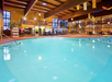 Indoor swimming pool area with lounge chairs, surrounded by two levels of balconies and large windows. Tables and chairs are on the poolside deck, with wooden beams on the ceiling.