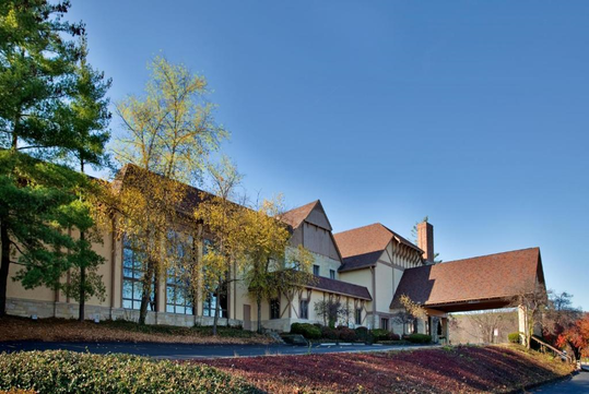 A large building with a sloped roof and Tudor-style architecture, surrounded by trees and bushes, with a clear blue sky above.