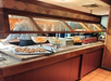 A buffet spread featuring pastries, bread rolls, and a variety of dishes with a person serving food in the background.