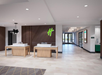 Hotel lobby with modern decor featuring two wooden reception desks, a patterned accent wall, green logo, and bright overhead lighting. Large windows in the background provide natural light.
