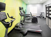 A small gym room with treadmills, an exercise bike, and shelving with white towels. A TV screen is mounted on the wall.