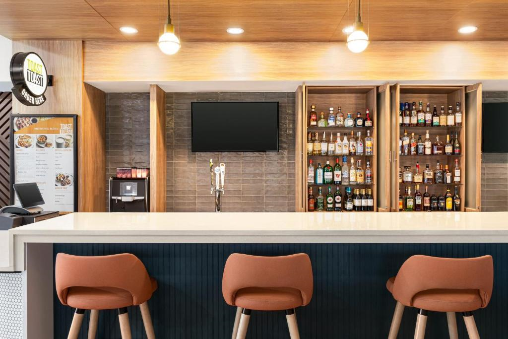 A modern bar area with three orange barstools, a countertop, and shelves stocked with various bottles. A menu is displayed on the left, and a TV is mounted centrally above the bar.