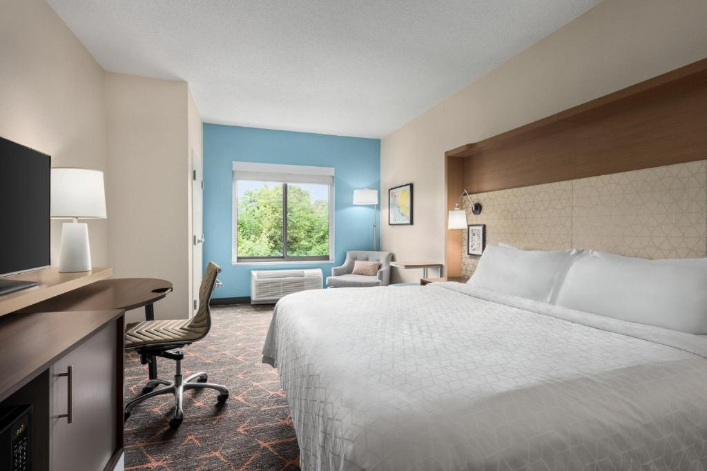 A hotel room with a large bed, desk, chair, and TV. There's a window with a view of trees and a small seating area with an armchair and lamp. The wall is partly painted blue.
