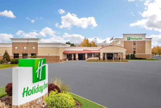 Holiday Inn hotel exterior with a large parking lot, brick facade, and prominent signage, set under a partly cloudy sky.