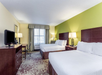 Hotel room with two double beds, a TV on a dresser, a desk with chair, and a window with curtains. Walls are green and beige, with carpeted flooring. Lamps are on bedside tables.