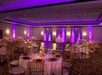 A decorated banquet hall with round tables, purple lighting, and an empty dance floor in the center.