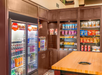 Snack shop with a refrigerated section for drinks on the left and shelves stocked with various snacks on the right. A wooden counter is in the foreground.
