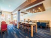 A well-lit dining area with wooden tables, red and blue upholstered chairs, and ceiling lights. Large windows allow natural light in.