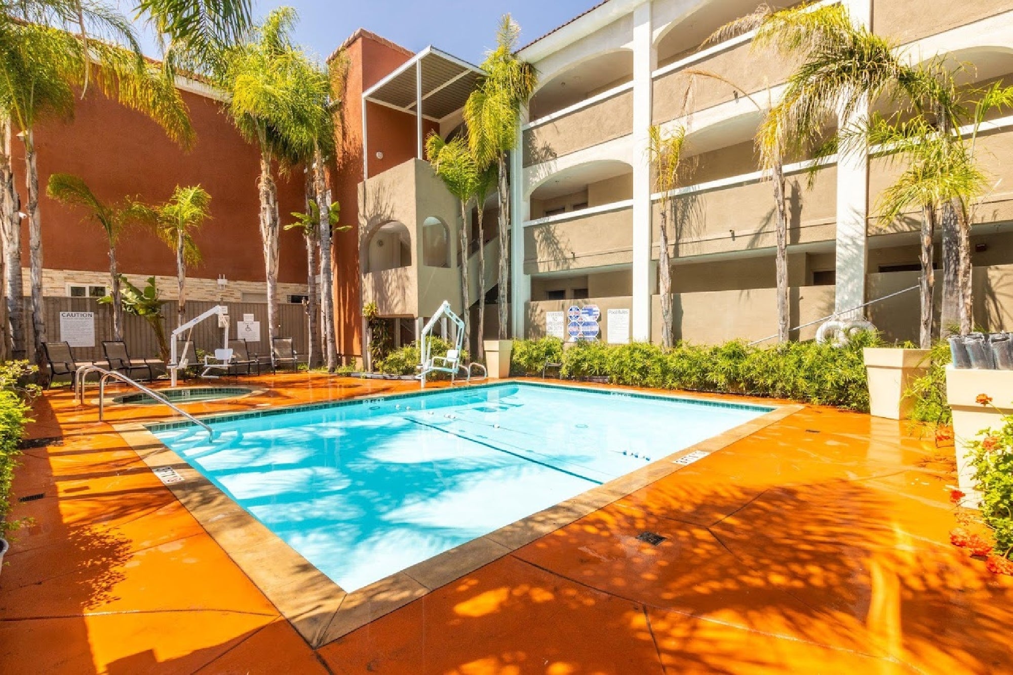 Outdoor courtyard with a small swimming pool surrounded by palm trees and lounge chairs, adjacent to a multi-story building.