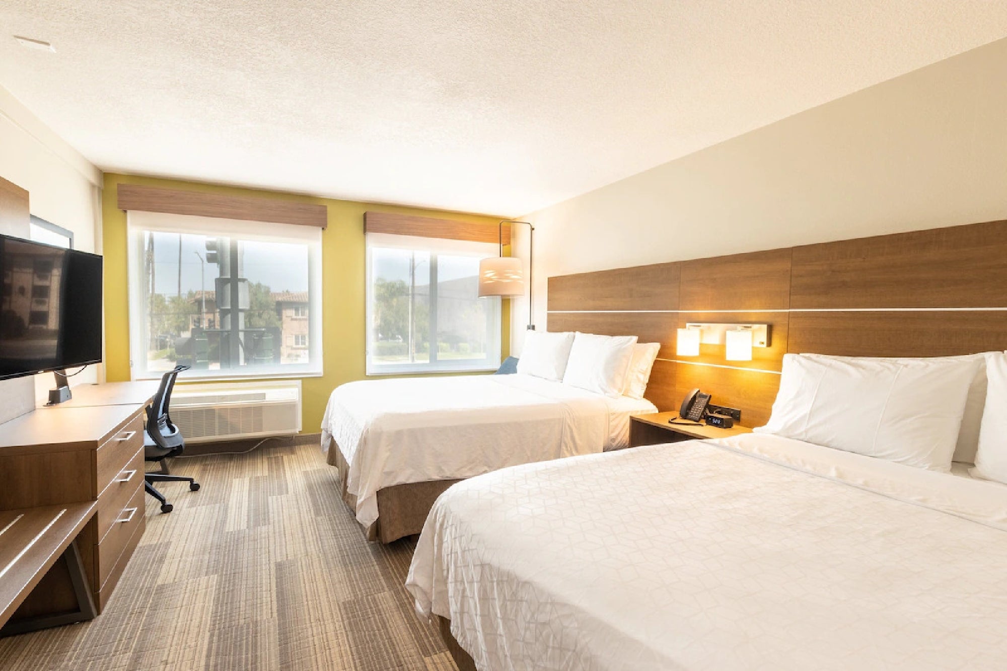 A hotel room with two double beds, a desk, a flat-screen TV, and large windows. The decor features light colors and wood accents.