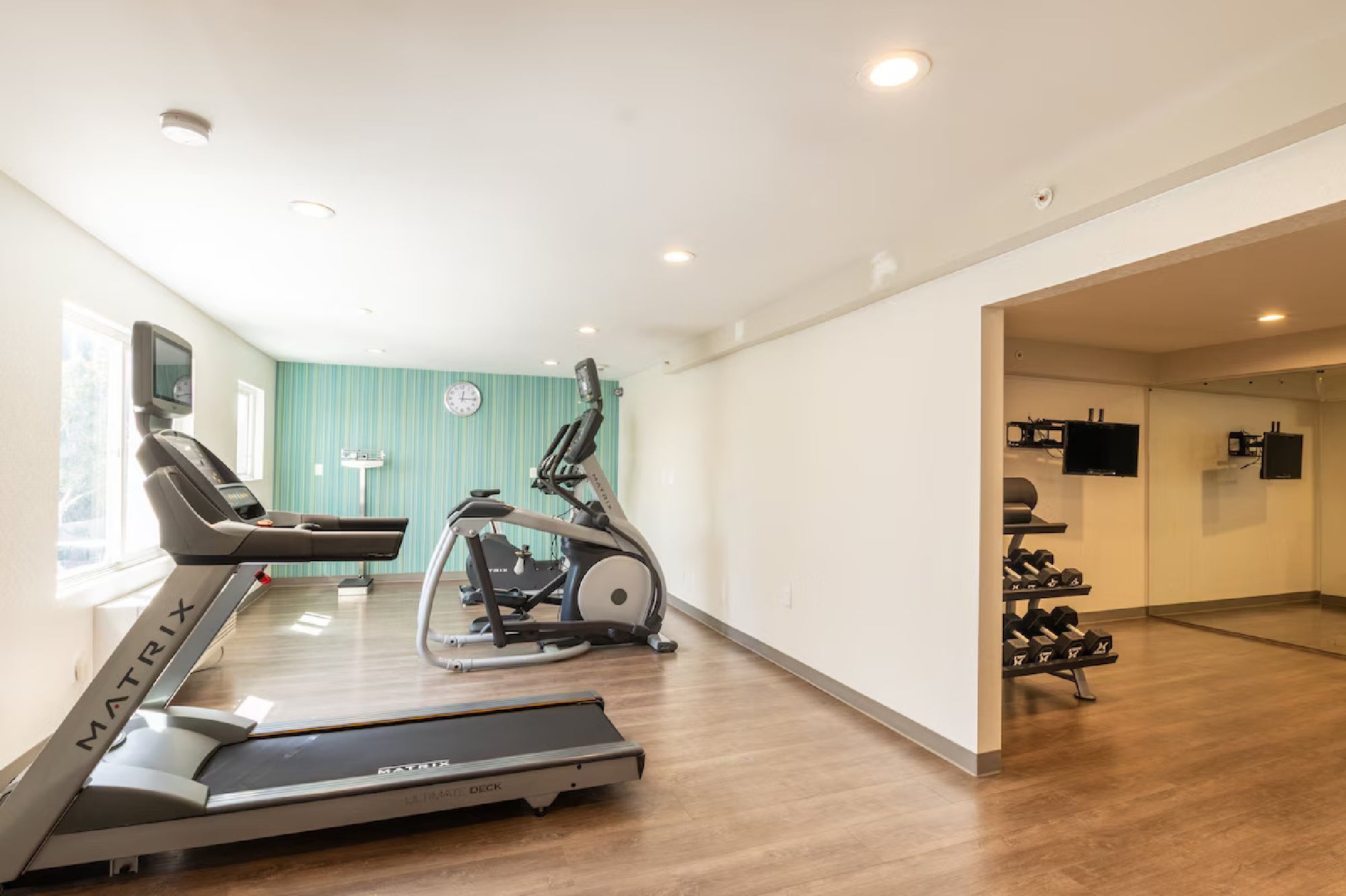 A small gym with two treadmills, an elliptical machine, a rack of dumbbells, and a wall clock. The room has wood flooring and a mirror on the right wall.