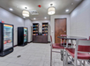 Small room with a table and four chairs, two beverage coolers, and shelves stocked with snacks. Ceiling lights illuminate the tiled floor and neutral-colored walls.