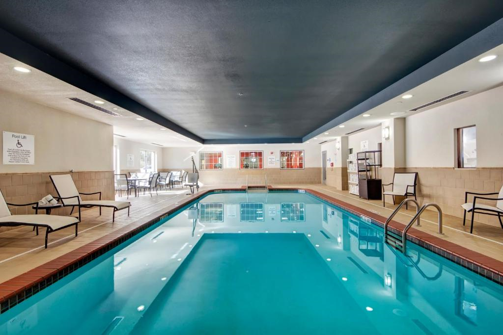 Indoor swimming pool area with blue water, surrounded by lounge chairs and tables. The walls are light-colored, and there are ceiling lights above.