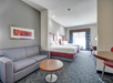 Hotel room with two beds, a sofa, round table, and a desk. Gray and red color scheme with patterned carpet, artwork on the wall, and a window with curtains in the background.